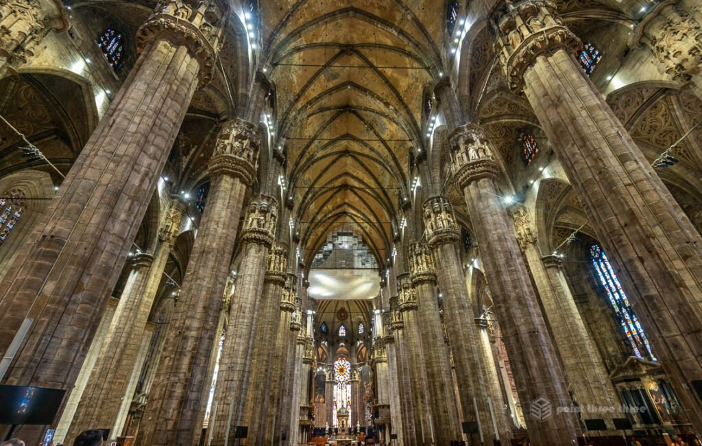 Interior view of the Duomo di Milano showing the high vaulted ceilings, massive stone columns, and sunlight streaming through stained glass windows.