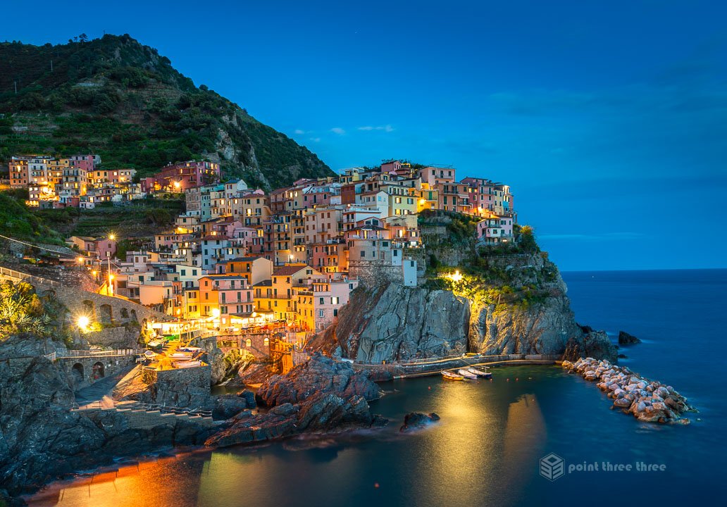 Manarola, Cinque Terre at night with glowing village lights reflecting on the ocean and a deep blue twilight sky.