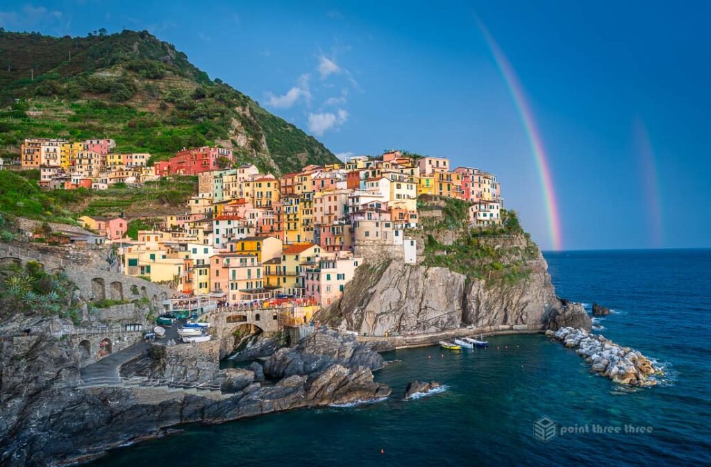 Panoramic view of Manarola village in Cinque Terre with a vibrant double rainbow over the Ligurian Sea and colorful cliffside houses.