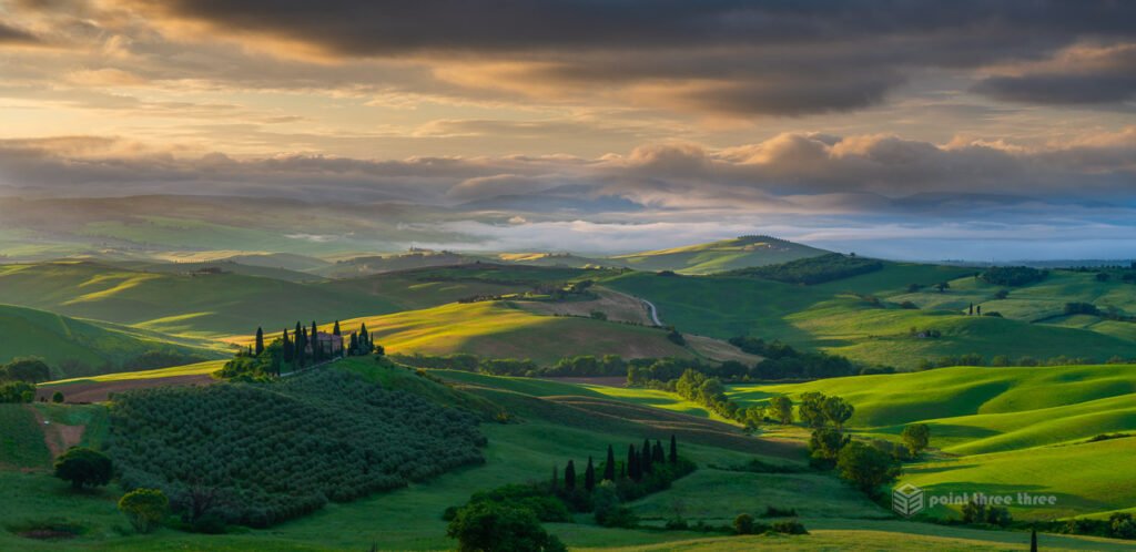 Sunrise at Podere Belvedere farmhouse in Val d'Orcia, Tuscany, featuring a cluster of cypress trees and rolling hills shrouded in morning mist.