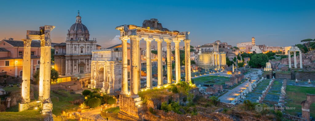 Panorama of the Roman Forum at blue hour with illuminated ancient ruins, including the Temple of Saturn and the Arch of Septimius Severus, under a twilight sky.