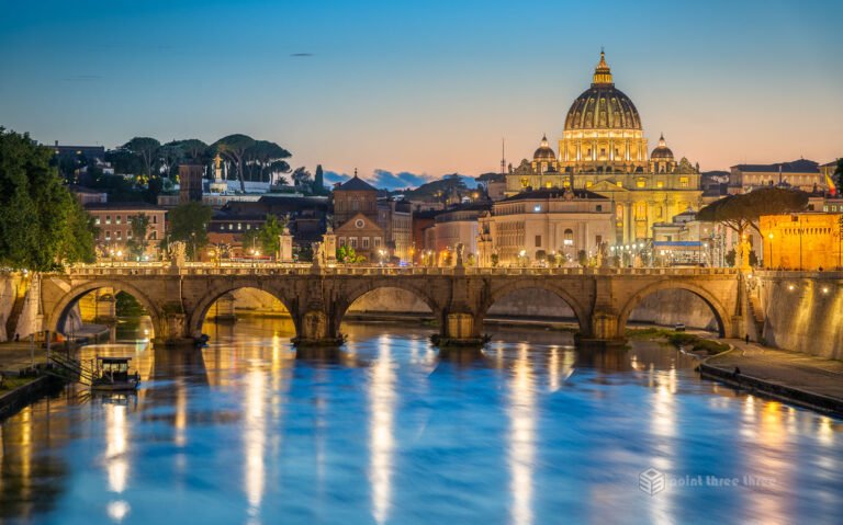 Blue hour view of St. Peter's Basilica dome and the Tiber River from Ponte Umberto I in Rome, with city lights reflecting on the water.