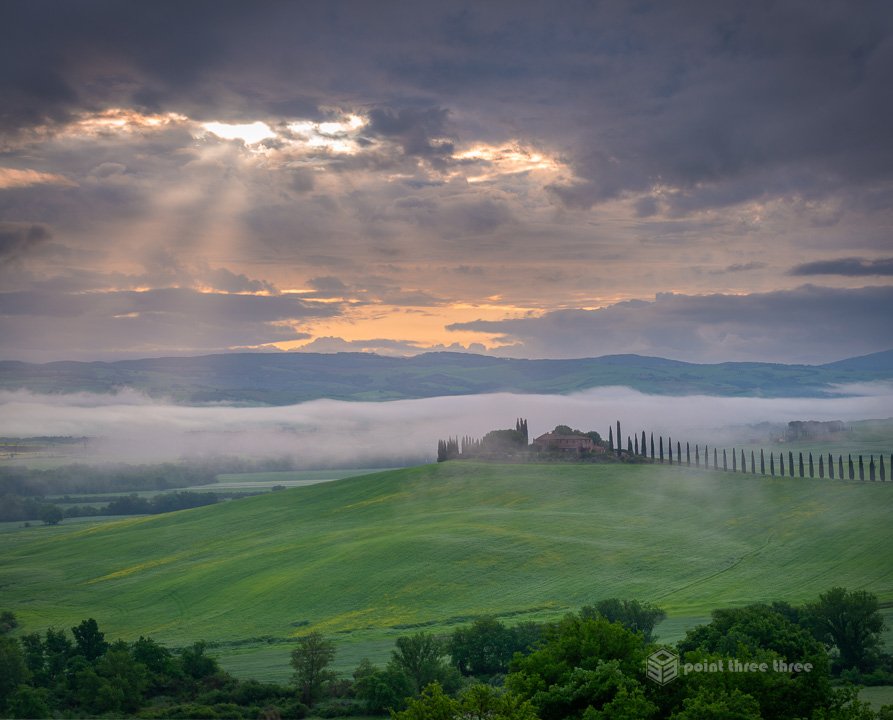 Sunrise with rays of light peaking through the clouds, a symmetrical row of cypress trees leading to a Tuscan farmhouse at Poggio Covili in Val d'Orcia, Italy.