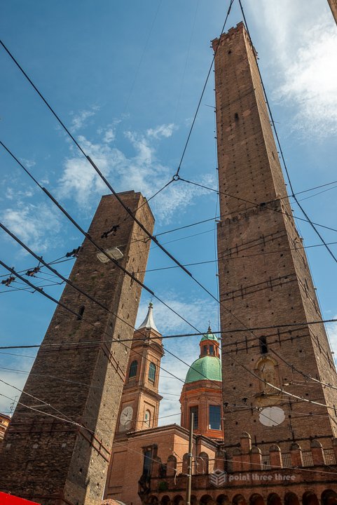 The Two Towers of Bologna, Asinelli and Garisenda, viewed from below against a blue sky with overhead tram wires.