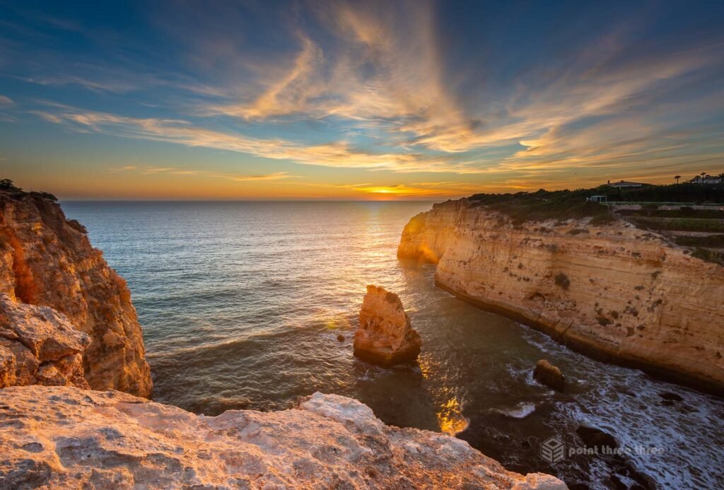 Dramatic sunset view from the clifftop above Praia de Vale Covo, Carvoeiro, Algarve, Portugal, with a lone sea stack glowing in golden light and a natural arch in the cliff face to the right.