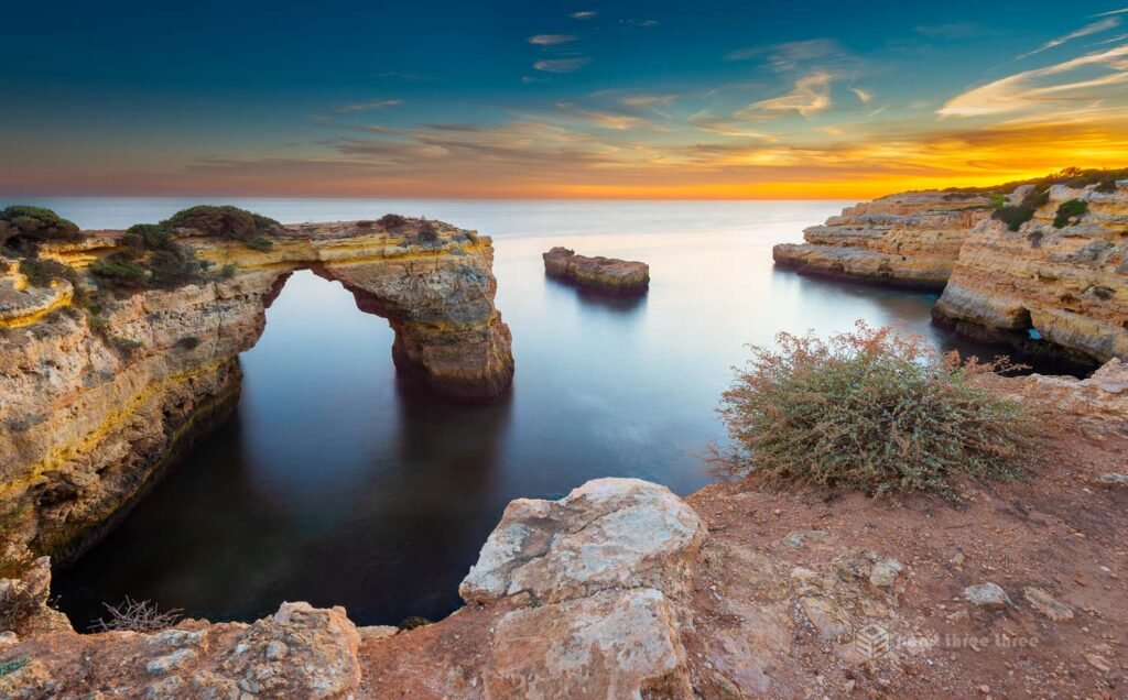 The Arco de Albandeira natural limestone sea arch at sunset, Algarve, Portugal, with mirror-calm water reflecting the colourful sky in a long exposure, and coastal scrub vegetation in the foreground.