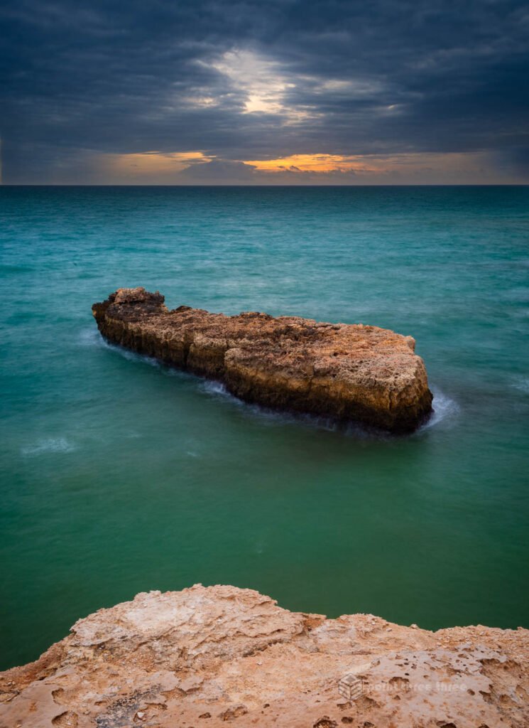 Long exposure seascape looking west from the top of the Arco de Albandeira natural limestone arch, Algarve, Portugal, with an isolated flat rock formation floating in jade-green Atlantic water under dramatic storm clouds and a glowing sunset horizon.