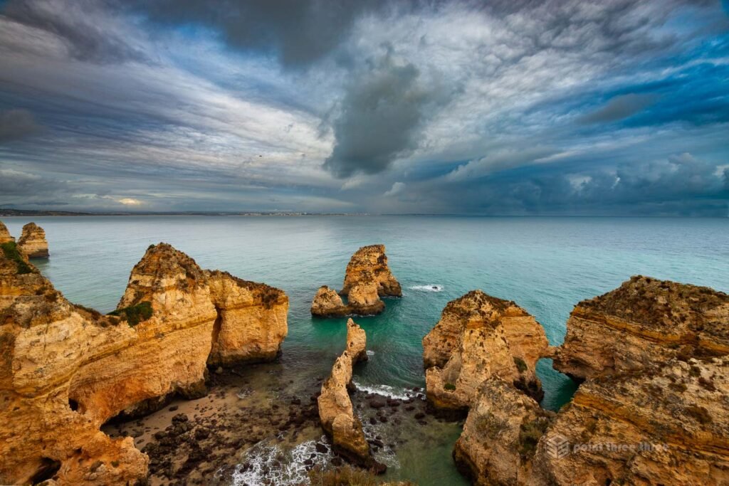 Wide angle view of the golden limestone sea stacks and arches of Ponta da Piedade under dramatic storm clouds, Lagos, Algarve, Portugal, with turquoise Atlantic water between the formations.