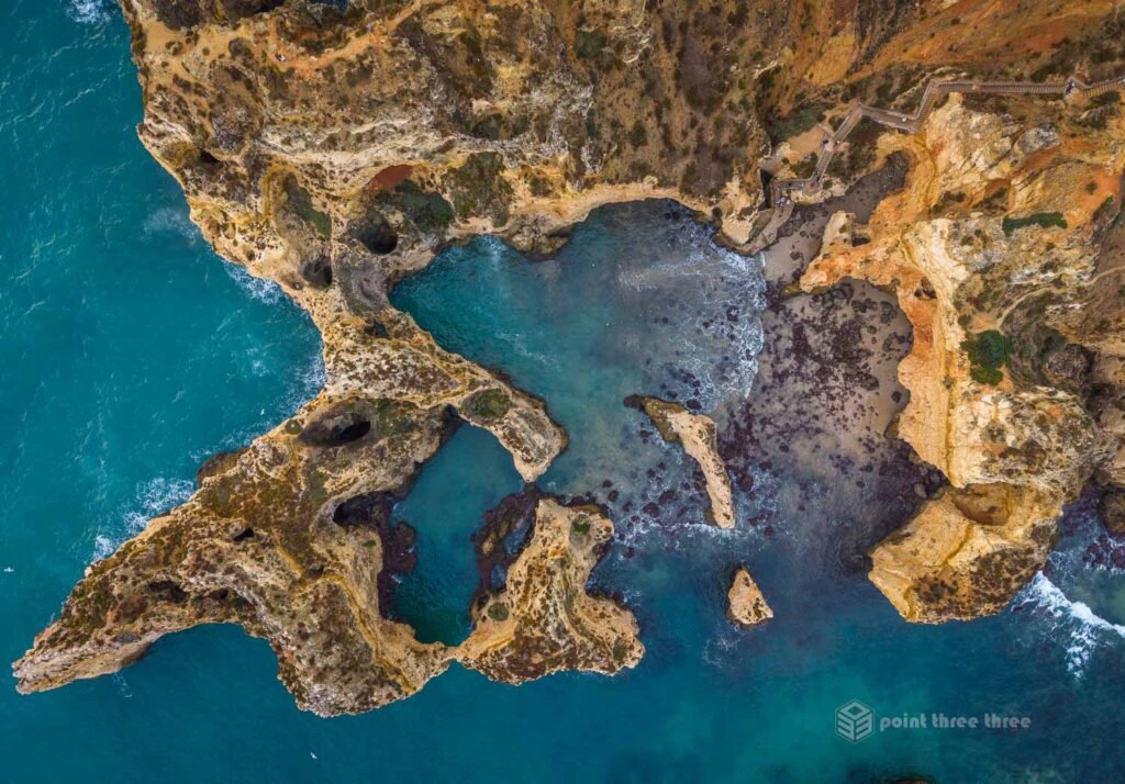 Straight-down aerial drone photograph of the Ponta da Piedade headland, Lagos, Algarve, Portugal, revealing the full labyrinth of limestone arches, grottos, sea caves and turquoise tidal pools carved into the golden cliffs, with the wooden staircase and viewing platform visible at upper right.