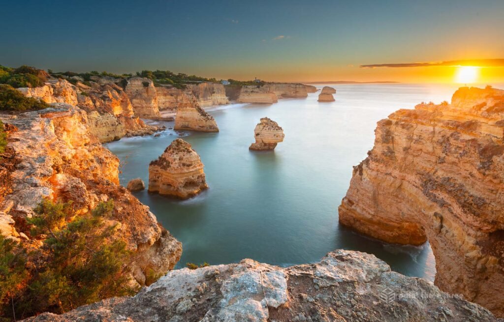 Panoramic long exposure sunrise view from the clifftops above Praia da Marinha, Algarve, Portugal, with multiple limestone sea stacks rising from smooth silky water in a sheltered cove, golden cliffs glowing in the first light and the sun rising over the Atlantic horizon.