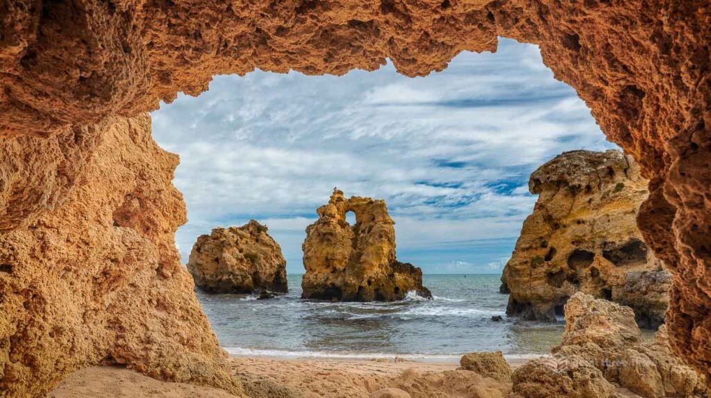The three limestone sea stacks of Praia dos Arrifes, Albufeira, Algarve, Portugal, photographed through a large natural cave arch in the cliff face, with golden sand beach and Atlantic waves visible beneath the dramatic rock frame.