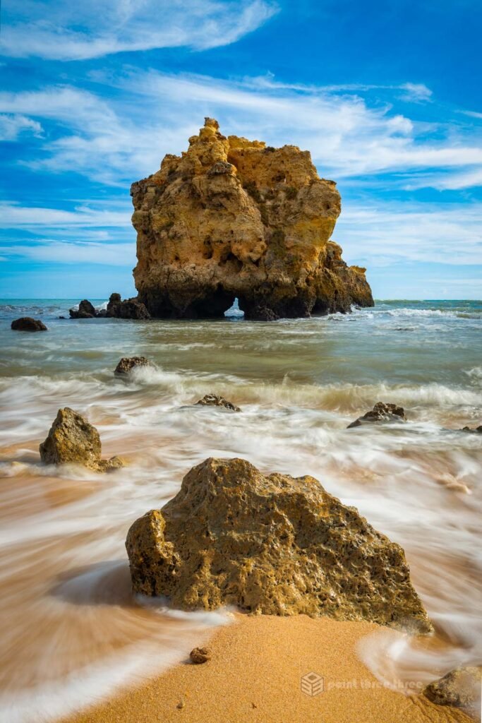Long exposure seascape of the iconic limestone sea stack at Praia dos Arrifes, Albufeira, Algarve, Portugal, with silky wave motion and rock foreground under blue sky.
